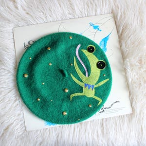 May include: A green beret featuring a cartoon frog character with a long pink tongue and black button eyes. The frog has a light green body and a blue collar. The beret is decorated with scattered gold-toned metal studs across the surface.