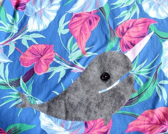 May include: A blue fabric featuring a vibrant tropical leaf pattern in shades of pink, white, and green. An appliqued gray narwhal with a textured finish is stitched onto the fabric, complete with a black button eye and a pale pink tusk.
