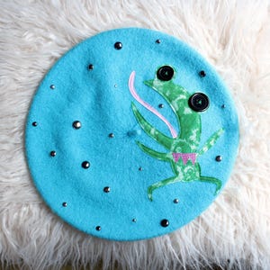 May include: A bright blue beret featuring a green frog appliqué with a long pink tongue. The frog has two black button eyes. The beret is decorated with scattered silver-toned metal studs of varying sizes across the surface of the fabric.