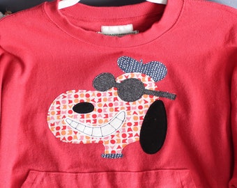 May include: A bright red long-sleeve sweatshirt featuring a large applique of a cartoon dog wearing dark glittery sunglasses and a blue patterned beret. The dog's face is filled with a geometric pattern of orange, pink, and white shapes.