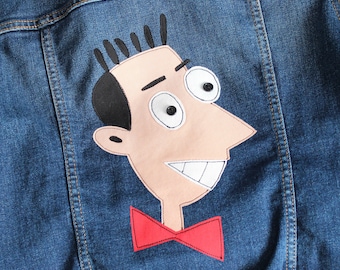 May include: Blue denim jacket with a cartoon character patch on the back. The patch features a light skin tone face with black hair, large eyes, and a red bow tie. The jacket has visible stitching.