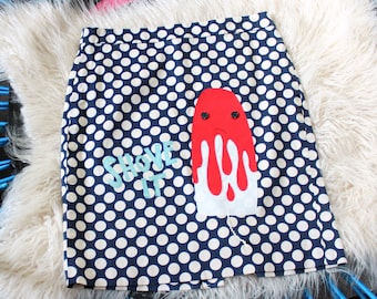 May include: A navy blue skirt with a white polka dot pattern featuring an appliqué of a red and white melting popsicle with two black button eyes and a sad mouth, alongside the embroidered light blue text that reads SHOVE IT.