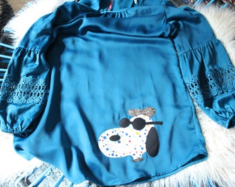 May include: A teal long-sleeve blouse featuring a round neckline and gathered sleeves with intricate lace detailing at the elbows. The front displays a white applique of a dog wearing black sunglasses and a glittery beret, accented with colorful dots.