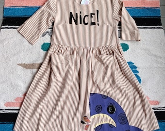 May include: A knee-length dress with brown and white vertical stripes, three-quarter sleeves, and a round neckline. The chest features the word NICE! in black letters. A purple patch with a black button eye and yellow teeth is on the skirt.