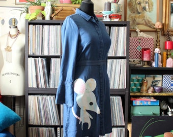 May include: A dark blue denim shirt dress featuring a long-sleeved design, a classic collar, and a button-down front. The lower portion of the dress displays a large, stylized white mouse graphic with a pink ear and a black button eye detail.