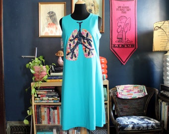 May include: A sleeveless turquoise dress featuring a central graphic of human lungs. The lung design is filled with a colorful floral pattern on a dark blue background with white speckles, creating a distinct contrast against the solid turquoise fabric.