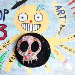 May include: A black felt beret featuring a central floral skull appliqué with pink, orange, and green petals. The beret is decorated with small silver star accents and is positioned over a yellow octopus illustration with text.