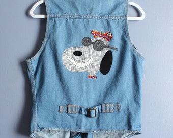 May include: A light blue denim vest featuring a large graphic of a cartoon dog wearing sunglasses and a colorful patterned hat. The vest includes a back cinch strap with a metal buckle and is displayed on a white plastic hanger.