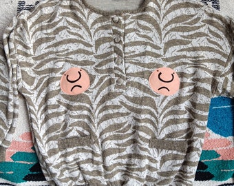 May include: A long-sleeved knit sweater featuring a beige and white zebra-like pattern, a button-up placket at the neckline, and two front pockets. Two circular peach-colored patches with black line drawings of frowning faces are sewn on the chest.