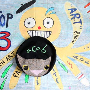 May include: A black beret featuring a light green embroidered text that reads "acab" above a brown patterned cat face applique. The cat face has two black button eyes, a pink triangular nose, and small white stitched details on the cheeks.