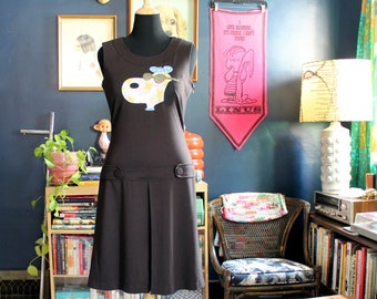 May include: A sleeveless dark brown dress featuring a graphic of a cartoon dog wearing a blue beret and gold glasses on the chest. The dress has a round neckline, a horizontal waistband with two decorative buttons, and a pleated skirt section.