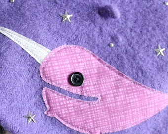 May include: A purple felt beret featuring a pink fabric narwhal appliqué with a black button eye and a white patterned horn. The beret is decorated with small silver star-shaped studs and tiny silver circular studs scattered across the surface.