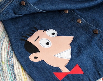 May include: A dark blue denim shirt featuring a large fabric appliqué of a cartoon character face with black hair, white eyes with black button pupils, and a wide grin. The character wears a red bowtie. The shirt has brown metal buttons.