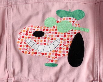 May include: A light pink denim jacket featuring a large applique of a cartoon dog character. The dog has a patterned face with red, orange, and pink geometric shapes, a black nose, a wide white smile, a black ear, and green glittery glasses.