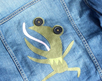 May include: A denim jacket with a green frog applique. The frog has a pink tongue and black button eyes.