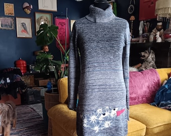 May include: A grey heathered knit sweater dress featuring a high turtleneck, long sleeves, and a straight hem. The lower front section displays a prominent graphic patch with a blue and white floral pattern and a solid red triangular accent.