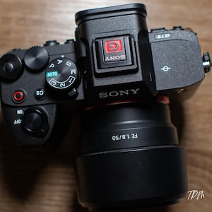 May include: Black Sony digital camera with a lens. The camera features dials and buttons, including an "AUTO" setting. The lens is marked "FE 1.8/50". The camera body has the Sony logo and the letters "G" and "C2".