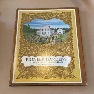 May include: A hardcover book titled "Pioneer Gardens" with a gold and cream-colored cover. The cover features an illustration of a white house and garden scene within a decorative floral frame. The book's title and author's name are printed in black.