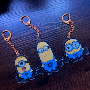 May include: Three acrylic keychains featuring Minion characters from the Despicable Me franchise. Each keychain has a yellow Minion figure wearing blue overalls, with orange key rings and chains. The Minions have different facial expressions.