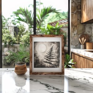 Puede incluir: Una ilustración en blanco y negro enmarcada de un helecho con una luna llena en el fondo. El marco es de madera y tiene un acabado rústico.