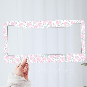 May include: A car number plate frame featuring a floral design of pink flowers and green leaves on a white backdrop. The frame is held by a hand. The text "Valley Vinyl Customs" is visible.