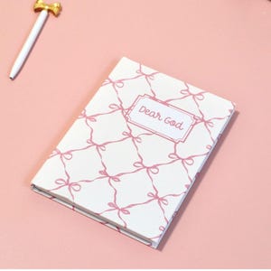 May include: A white notebook with a pink bow pattern and the words "Dear God" on the cover. A gold and white pen is next to the notebook on a pink surface. Flowers are in the background.