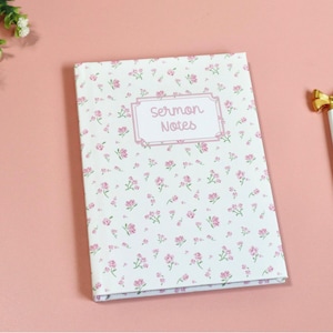 May include: A floral-patterned notebook with the words "Sermon Notes" in a pink frame, next to a white pen with a gold bow-shaped top. The notebook and pen rest on a pink surface, with flowers in the upper left corner.