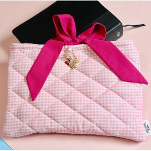 Pink gingham book sleeve cover quilted with pink ties for closure fashioned into a coquette bow. Inside of sleeve is lined with pink fabric and includes an internal pocket to hold small accessories. The outside of the book protector includes a a gold charm ring with a cherry charm, coquette bow charm, a cross charm and a pearl charm. A custom initial charm can be added for personalization.