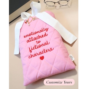 Light pink quilted book sleeve with red embroidered lettering and white bow closure, reading “emotionally attached to fictional characters.” The sleeve is styled alongside a thick black novel and a pair of round reading glasses, creating a cozy, bookish setup ideal for avid readers and fictional world lovers.