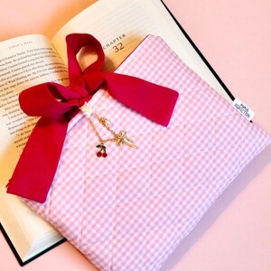 May include: A pink and white gingham book sleeve with a red ribbon and a gold cherry charm necklace. The book sleeve is open, revealing pages from a book. The text "BUDGET FRIENDLY GIFTS FOR BOOK LOVERS" is at the bottom.