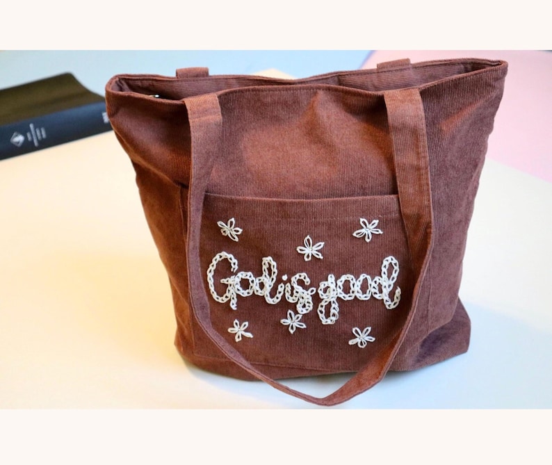 Handmade corduroy Christian tote bag featuring “God is Good” beautifully hand embroidered in textured script. Designed with a large front pocket, secure zipper closure, and multiple interior compartments, perfect for carrying your Bible, notebook, or daily essentials. Ideal as a faith-based gift for women, a church tote, or Bible study bag. A durable, stylish, and meaningful Christian accessory for everyday inspiration.