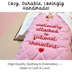 Close-up of a light pink quilted book sleeve on a vibrant blue background, highlighting the detailed stitching and bold red embroidered phrase “emotionally attached to fictional characters.” White ties add a delicate closure. On-screen text overlay reads: “cozy. durable. lovingly handmade. high quality quilting and embroidery – made to love and last.”