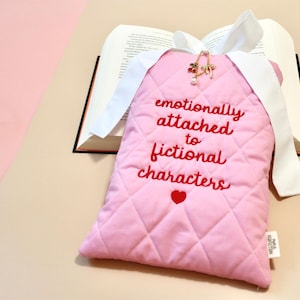 Handmade quilted book sleeve featuring the embroidered phrase "Emotionally Attached to Fictional Characters" with a heart design below in red thread on soft pink fabric.