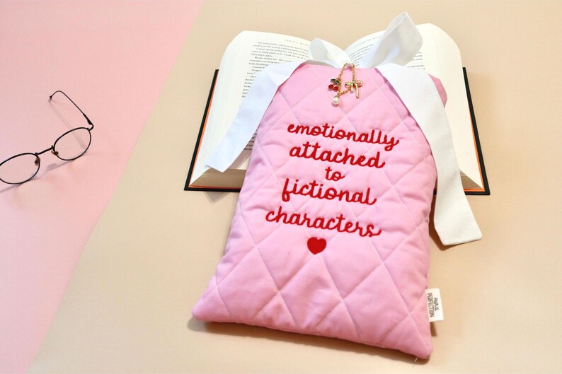 Handmade quilted book sleeve featuring the embroidered phrase "Emotionally Attached to Fictional Characters" with a heart design below in red thread on soft pink fabric.