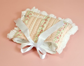 May include: A baby bonnet with a floral pattern, quilted design, and the name "Aubrey" embroidered on it. The bonnet has a pink and cream colour scheme, white lace trim, and a white satin ribbon tied in a bow.
