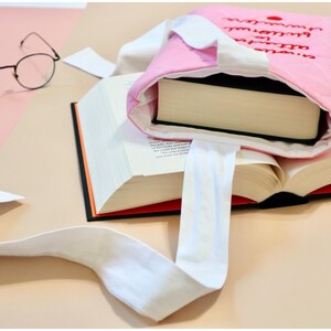 May include: A pink quilted fabric book sleeve with white fabric handles and black embroidered text on the front. The sleeve contains a thick hardcover book, and a large open book lies underneath it, accompanied by a pair of thin-rimmed round glasses.