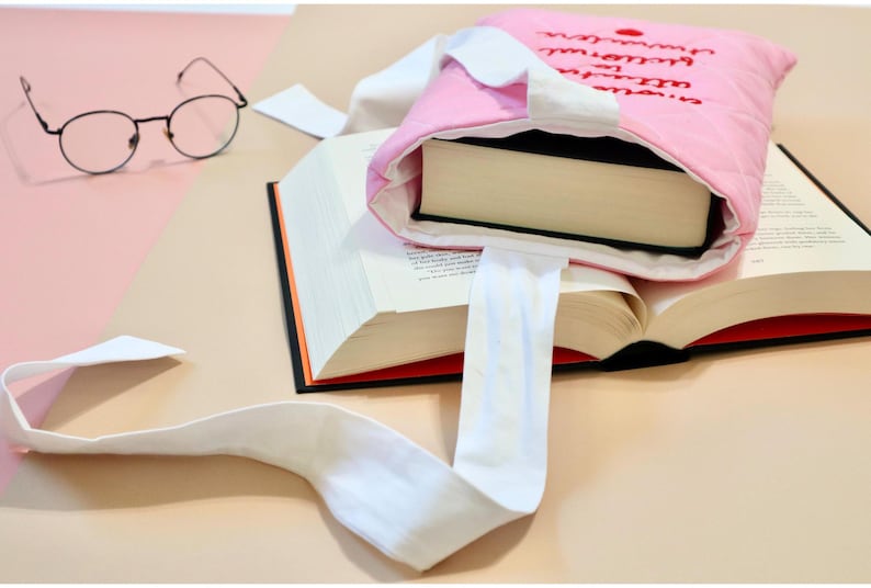May include: A pink quilted fabric book sleeve with white fabric handles and black embroidered text on the front. The sleeve contains a thick hardcover book, and a large open book lies underneath it, accompanied by a pair of thin-rimmed round glasses.