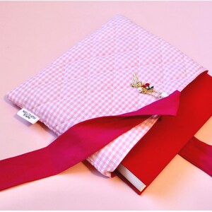 cloth book cover pink lying open on a pink background and displays a red hardcover novel half way inserted into the book sleeve pouch. also visible is the charm bracelet that includes a cherry, coquette bow and a pearl charm.