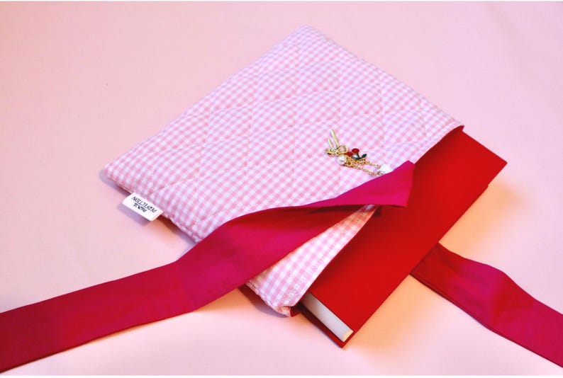 cloth book cover pink lying open on a pink background and displays a red hardcover novel half way inserted into the book sleeve pouch. also visible is the charm bracelet that includes a cherry, coquette bow and a pearl charm.