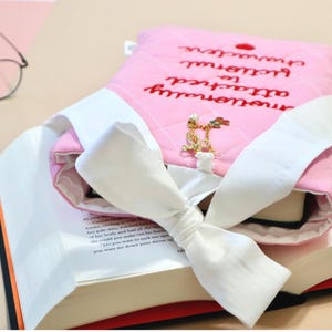 Pink quilted book sleeve for book lovers with red embroidered text reading "Emotionally Attached to Fictional Characters" and a heart accent, designed to protect books and e-readers.