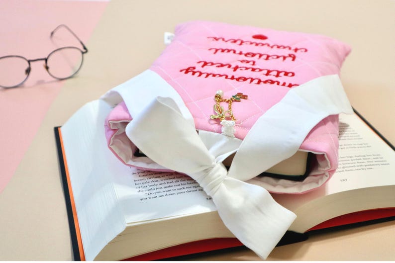 Pink quilted book sleeve for book lovers with red embroidered text reading "Emotionally Attached to Fictional Characters" and a heart accent, designed to protect books and e-readers.