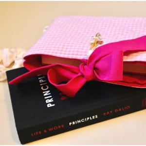 Pink Bible Cover Quilted Custom Bible Book Cover Book Sleeve With Bow Book Bag For Mom Book Lover Gift For Teen Daughter Birthday Present For Goddaughter baptism gift for best friend book lover. a pink gingham book protector with dark pink coquette bow tie closure sitting atop of a black hardcover book
