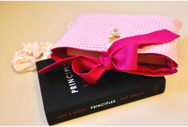 Pink Bible Cover Quilted Custom Bible Book Cover Book Sleeve With Bow Book Bag For Mom Book Lover Gift For Teen Daughter Birthday Present For Goddaughter baptism gift for best friend book lover. a pink gingham book protector with dark pink coquette bow tie closure sitting atop of a black hardcover book
