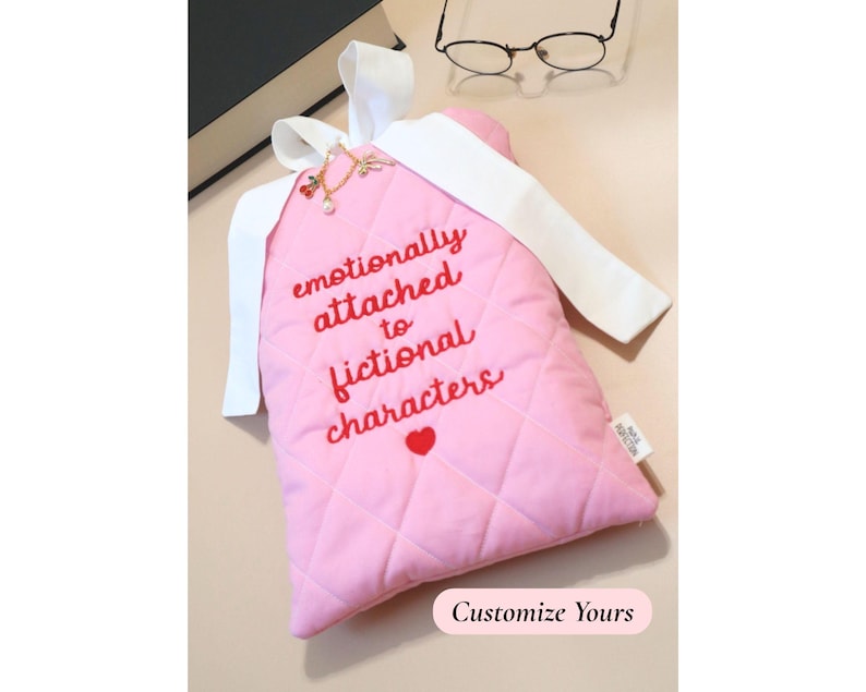 Light pink quilted book sleeve with red embroidered lettering and white bow closure, reading “emotionally attached to fictional characters.” The sleeve is styled alongside a thick black novel and a pair of round reading glasses, creating a cozy, bookish setup ideal for avid readers and fictional world lovers.