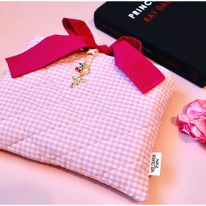handmade quilted bow bible cover pink plaid fabric book cover with coquette bow closure. Book sleeve sits to the side of a black hardcover book and a pink satin scrunchie lies on the table next to them. The pink bible sleeve would be the perfect gift for a teenage girl baptism or communion gift, or a unique girlie gift for a bookish girl summer reading lover.