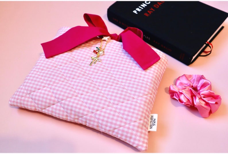 handmade quilted bow bible cover pink plaid fabric book cover with coquette bow closure. Book sleeve sits to the side of a black hardcover book and a pink satin scrunchie lies on the table next to them. The pink bible sleeve would be the perfect gift for a teenage girl baptism or communion gift, or a unique girlie gift for a bookish girl summer reading lover.