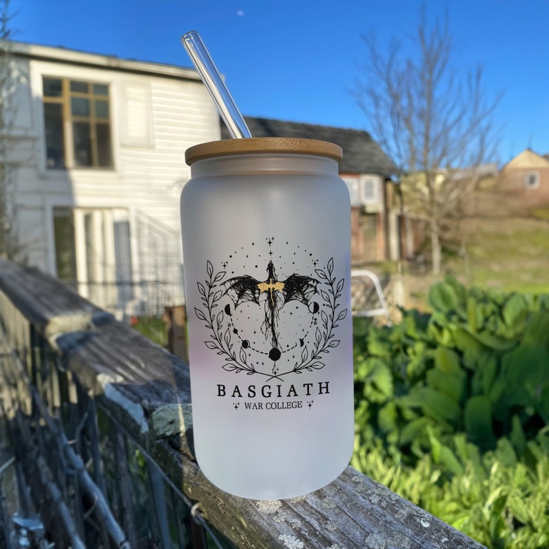 May include: A frosted glass water bottle with a bamboo lid and a black and gold design of a dragon with stars and moons. The bottle has the text "BASGIATH WAR COLLEGE" printed on it.