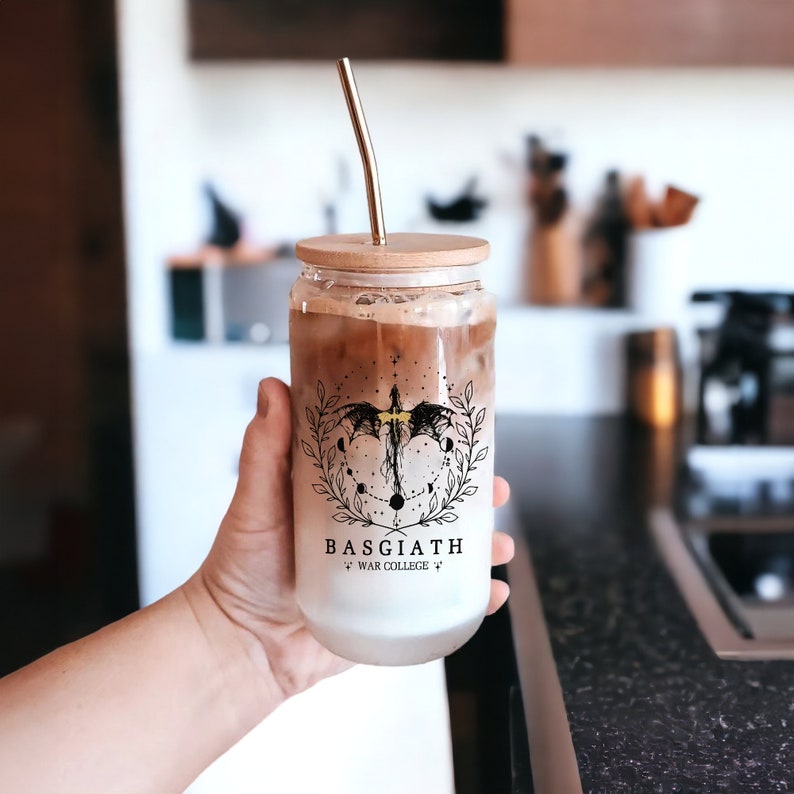 May include: A clear glass tumbler with a black and white graphic of a dragon with a gold accent. The tumbler has a wooden lid and a straw. The tumbler has the text "Basgiath War College" printed on it.