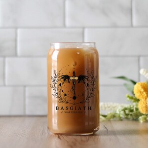 May include: A clear glass can with a black and gold design featuring a dragon, stars, and the text "BASGIATH WAR COLLEGE". The can is filled with a light brown liquid.
