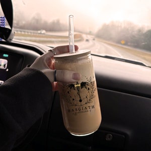 May include: A clear glass tumbler with a wooden lid and a black and gold design. The design features a winged creature with a crescent moon and stars. The text on the tumbler reads "Basgiath War College".